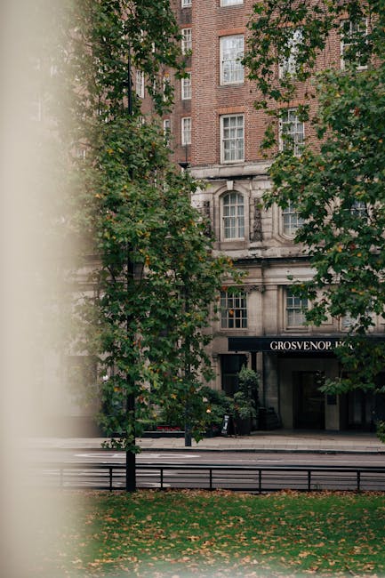 A view of a multi-storey brick building with large windows, partly obscured by green trees with broad leaves, situated on a street near Grosvenor Square in Mayfair. The building's entrance features a black awning with white lettering that reads 'GROSVERNOR' and is partially visible behind the trees. In the foreground, a black metal fence runs parallel to the pavement, which is covered with fallen leaves, indicating an autumn setting. This scene captures the exterior environment where house removals and furniture transport activities might take place, with the natural setting and urban architecture providing context for relocation services by Removal Companies Mayfair. The lighting appears natural, with daylight filtering through the foliage, creating a calm and professional atmosphere suitable for describing aspects of home relocation or moving logistics.