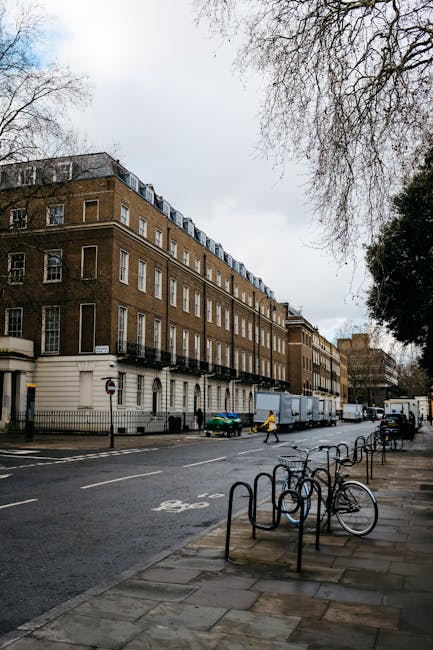 The image depicts a street scene outside a multi-storey Victorian-style building with a brown brick facade and white window frames, located near Grosvenor Square in Mayfair. Several large cardboard boxes and moving cartons are stacked on the sidewalk adjacent to a vehicle, likely part of a house removal or furniture transport process. A person in bright clothing is visible, walking near the boxes, while another individual is engaged in loading or unloading items from a light-colored van parked on the street. The vehicle is positioned close to the building's entrance, suggesting active packing or unpacking. In the foreground, bicycle racks with a single bike are present on the sidewalk, alongside metal fencing separating the pedestrian area from the road. Overhead, leafless trees partially cover the cloudy sky, creating a typical urban environment for home relocation services. This scene illustrates a professional moving operation taking place in an urban residential area, aligned with the services offered by [COMPANY_NAME] for efficient house removals near Grosvenor Square, as detailed on [PAGE_TITLE].