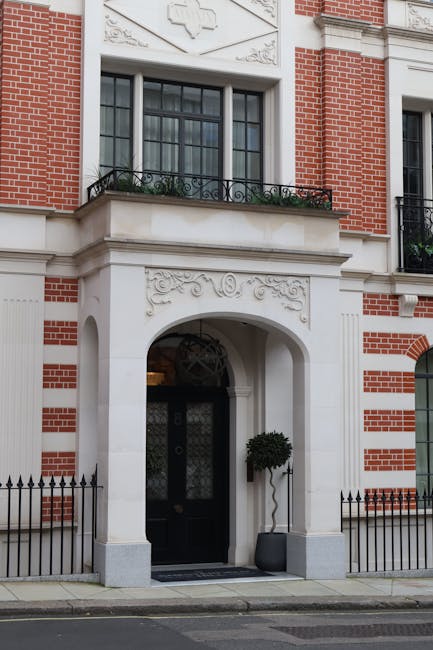 A view of a multi-storey brick building with large windows, partly obscured by green trees with broad leaves, situated on a street near Grosvenor Square in Mayfair. The building's entrance features a black awning with white lettering that reads 'GROSVERNOR' and is partially visible behind the trees. In the foreground, a black metal fence runs parallel to the pavement, which is covered with fallen leaves, indicating an autumn setting. This scene captures the exterior environment where house removals and furniture transport activities might take place, with the natural setting and urban architecture providing context for relocation services by Removal Companies Mayfair. The lighting appears natural, with daylight filtering through the foliage, creating a calm and professional atmosphere suitable for describing aspects of home relocation or moving logistics.